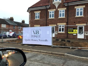 A white printed Heras Fence Cover on a heras fence panel at a construction site with 2 empty heras fence panels on each side of it.